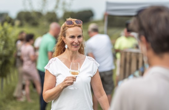 Vrouw met een glas wijn, lachend op een buitenfeest in de natuur. (vergroot foto)